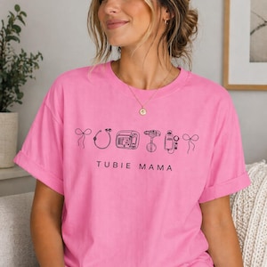 以下が含まれることがあります： 鮮やかなピンク色のTシャツ。黒字で「TUBIE MAMA」と書かれています。その上に、医療機器の黒い線画が描かれています。袖はロールアップされ、ゆったりとしたフィット感です。