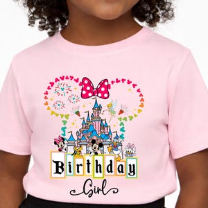May include: Pink t-shirt featuring a colorful graphic of a castle, fireworks, and characters. The design includes a polka-dot bow and the words "Birthday Girl" in a decorative font. A fun and festive design for a birthday celebration.