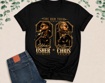 Peut inclure: Un t-shirt noir avec une impression graphique intitulée THE R&B TOUR 2026. Le design comprend des portraits d'Usher et de Chris Brown dans des cadres dorés, avec leurs noms imprimés en dessous en lettres dorées, accompagné d'un jean bleu.