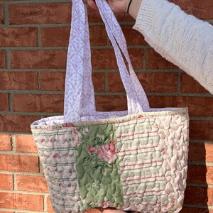 May include: A quilted tote bag with floral and striped patterns in shades of pink, green, and white. The bag features a patchwork design with a green central panel adorned with pink roses. The handles are a light pink with a repeating pattern.