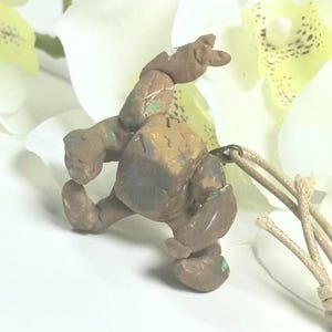 Puede incluir: Una pequeña figura de arcilla marrón y texturizada con forma de criatura robusta, extremidades redondeadas y torso cuadrado. La figura presenta sutiles marcas verdes y gris oscuro en su superficie y está unida a un cordón de color beige claro.