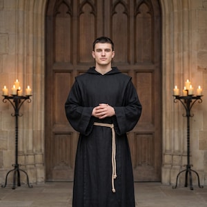 May include: A person wearing a black hooded robe with a beige rope belt, standing in front of a large wooden door. Two black wrought iron candle holders with lit candles flank the person.