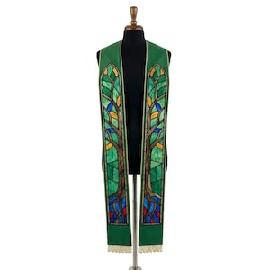 May include: A green liturgical stole featuring a stained glass window pattern with brown tree trunks, green leaves, and accents of blue, yellow, and red. The garment has a pointed top, straight vertical panels, and a fringe trim at the bottom edge.