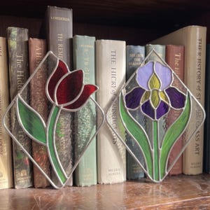 May include: Two diamond-shaped stained glass panels featuring floral designs. The left panel displays a deep red tulip with green leaves. The right panel depicts a purple and yellow iris with green leaves. Both pieces are framed in silver metal.