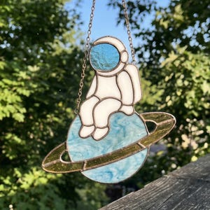 May include: A stained glass astronaut figure, in a white spacesuit, sits on a blue planet with a gold ring. The astronaut's helmet has a blue reflective surface. The piece hangs from a chain.