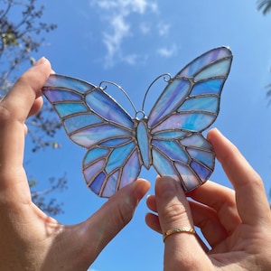 Puede incluir: Una mariposa de vidriera en tonos azules, morados y blancos, sostenida contra un cielo azul claro. Las alas están segmentadas y los bordes están delineados con un material metálico. Un artículo decorativo.