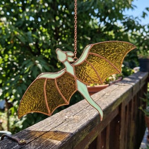Pode incluir: Um apanhador de sol de dragão em vitral com uma corrente de cobre. O dragão apresenta um corpo verde claro e asas texturizadas amarelo dourado. O apanhador de sol está pendurado ao ar livre contra um fundo verde desfocado.