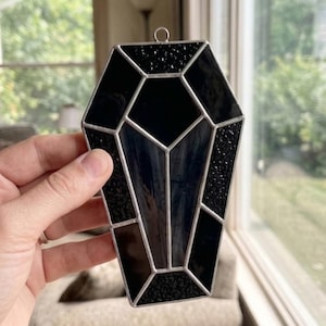 May include: A coffin-shaped stained glass suncatcher featuring a geometric arrangement of black glass panels with varying textures, including smooth and pebbled surfaces, held together by silver-toned metal solder lines and a small top hanging loop.