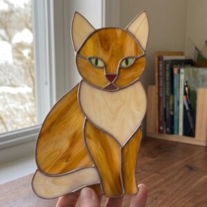 May include: A stained glass cat figurine in shades of orange, cream, and brown. The cat has green eyes and a red nose. The figurine is sitting upright with its ears perked up. The glass is outlined with a dark brown border.