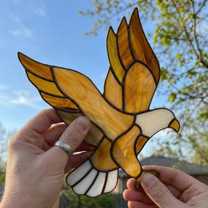 May include: A stained glass eagle in flight, with amber, gold, and white panels. The eagle's wings are outstretched, displaying the detailed glasswork. The artwork is held against a blue sky and green foliage. The piece is a decorative item.
