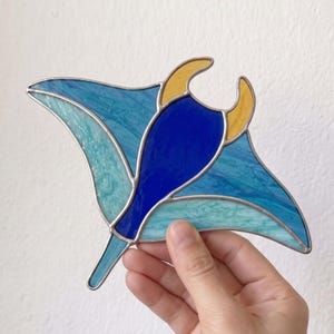 May include: A stained glass manta ray suncatcher featuring a deep blue central body, light blue and turquoise marbled wings, and two yellow horn-like protrusions at the top, all held together by thin silver-coloured metal solder lines.
