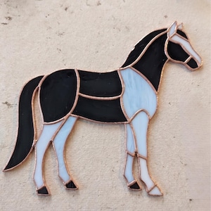Op de afbeelding: Een gebrandschilderde paardenfiguur in een staande pose. Het paard heeft een zwart lichaam, een wit gezicht en witte benen, en lichtblauwe accenten. De glazen stukken zijn omlijnd met koperkleurig metaal.