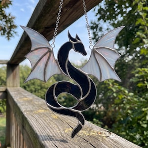 May include: A stained glass suncatcher shaped like a dragon with a black body and iridescent white wings. The dragon is suspended by a silver metal chain, featuring a curved tail and detailed wings constructed with thin metal solder lines.