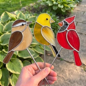 Könnte beinhalten: Drei Buntglas-Vogelornamente auf Metallstäben. Ein Vogel ist braun und beige, einer ist leuchtend gelb und einer ist kräftig rot mit einer schwarzen Gesichtsmaske. Jeder Vogel besteht aus einzelnen Glasteilen, die mit dünnen Metallnähten verbunden sind.
