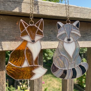 May include: Two stained glass suncatchers hanging from metal chains. One is a fox in shades of amber, orange, and white. The other is a raccoon in shades of grey, black, and white. Both feature geometric glass segments joined by dark metal solder lines.