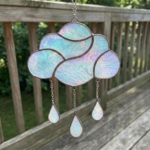 Op de afbeelding: Een glas-in-lood zonnevanger in de vorm van een wolk met een iriserende, parelmoerachtige afwerking. De wolk is opgebouwd uit meerdere segmenten verbonden door donkere metalen soldeerlijnen en heeft drie bungelende, traanvormige regendruppels van glas.
