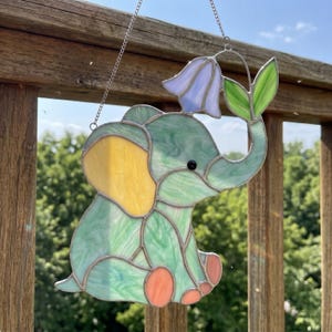 Puede incluir: Atrapasoles de elefante de vidriera en tonos verdes, amarillos y naranjas. El elefante sostiene una flor azul con hojas verdes. El atrapasoles cuelga de una cadena plateada, perfecto para una habitación infantil.