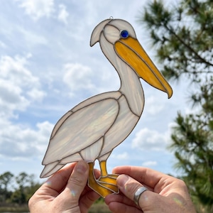 May include: A stained glass pelican suncatcher. The bird features a white body, yellow beak and feet, and a blue eye. The pelican is held up against a backdrop of a cloudy sky and green foliage.
