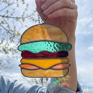 May include: Stained glass hamburger suncatcher with a chain for hanging. The burger features layers of yellow, red, green, and orange glass, resembling cheese, a patty, lettuce, and a bun. The glass has a textured appearance.