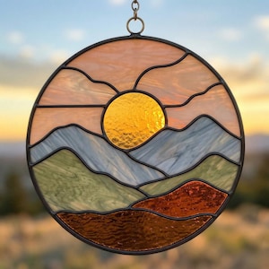 May include: Round stained glass art piece depicting a sunset over mountains. The sun is a bright gold circle, with layers of mountains in shades of blue, green, and brown. The sky is a mix of peach and orange hues, all framed in a black border.