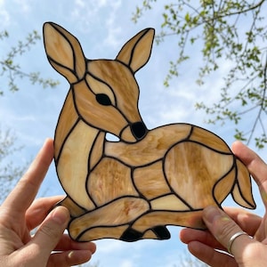 Könnte beinhalten: Ein Glasfenster-Reh in Braun- und Cremetönen mit schwarzen Umrissen. Das Reh befindet sich in einer Ruheposition, mit gedrehtem Kopf und angezogenen Beinen. Das Kunstwerk wird vor einem blauen Himmel mit Ästen im Hintergrund gehalten.