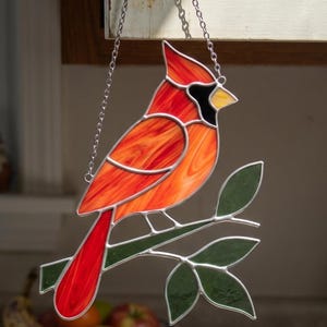 May include: Stained glass cardinal bird suncatcher. The bird is mainly red and orange with black and yellow details. It is perched on a green branch with green leaves. The suncatcher hangs from a silver chain.