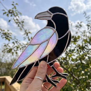 May include: A stained glass raven suncatcher featuring opaque black glass for the head and tail, contrasted with iridescent, opalescent glass on the wing that reflects soft pastel colours. The bird is constructed with thin, silver-toned metal solder lines.