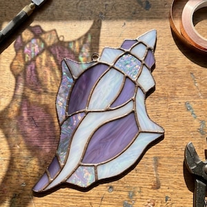 Könnte beinhalten: Handgefertigte Glas-Seeschnecke in Lila, Weiß und schillerndem Glas, mit Kupferfolienkanten. Das dekorative Kunstwerk ist so konzipiert, dass es das Licht einfängt. Ideal für Küsten- oder Stranddekoration.