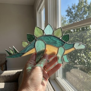 May include: A stained glass Stegosaurus dinosaur in shades of green, yellow, and blue. The dinosaur has a spiky back and is held up to a window. The glass is outlined with a silver metal.