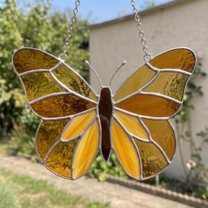May include: A stained glass butterfly suncatcher featuring amber, golden yellow, and dark brown glass segments held together by silver metal solder lines. The butterfly has two thin silver antennae and is suspended by a silver metal chain.