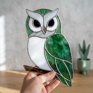 Könnte beinhalten: Glasmalerei-Eulenfigur mit grün-weißem Design. Die Eule hat große schwarze Augen, ein weißes Gesicht und grüne Flügel. Sie sitzt auf einem braunen Ast. Das Glas hat eine strukturierte Oberfläche.
