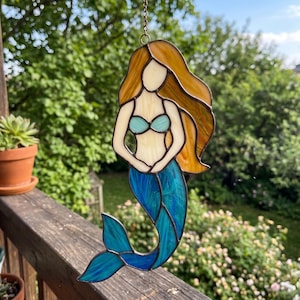 Pode incluir: Apanhador de sol de sereia em vitral com top de biquíni turquesa e cauda azul. A sereia tem cabelos longos e dourados esvoaçantes e está pendurada em uma corrente. O apanhador de sol é exibido ao ar livre com um fundo verde desfocado.