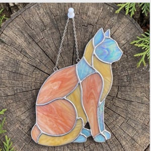 May include: Stained glass cat suncatcher in a sitting pose. The cat features a mix of orange, yellow, and blue glass pieces, with a silver chain for hanging. The cat is set against a rustic wooden background.