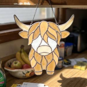 May include: Stained glass Highland cow suncatcher with a chain. The cow features amber, cream, and white glass. The artwork is designed with a detailed, segmented pattern. It is suspended in front of a window, catching the light.