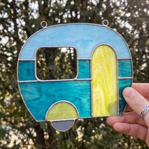 May include: A stained glass camper with a light blue roof, teal body, and yellow door. The camper has a window and a gray wheel. The camper is held up by a silver ring.