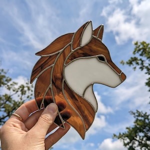 Peut inclure: Une œuvre d'art en vitrail représentant une tête de loup. L'œuvre présente des sections de verre marron et blanc, avec un contour en métal argenté. L'œuvre est tenue devant un ciel bleu avec des arbres en arrière-plan.