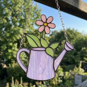 May include: A stained glass hanging featuring a lavender watering can with a pink flower and green leaves. The flower has a yellow center. The piece is suspended by a silver chain, perfect for garden or home decor.