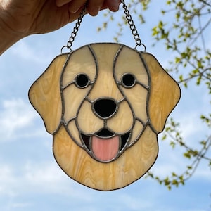 Puede incluir: Cabeza de perro de vidriera con una cadena para colgar. La cara del perro es de color amarillo claro con ojos negros y una lengua rosa. El vidrio está enmarcado con metal oscuro. El fondo es un cielo azul con algunas ramas de árboles.