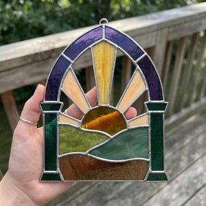 Könnte beinhalten: Ein bogenförmiger Sonnenfänger aus Buntglas mit einem zentralen Sonnenstrahlenmotiv in Gelb und Bernstein über hügeligen Landschaften in Grün- und Brauntönen. Das Stück ist oben mit dunkelviolettem Glas und seitlich mit grünen Säulen gerahmt.