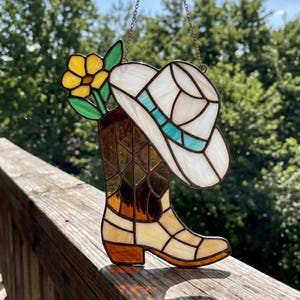 May include: Stained glass art piece featuring a cowboy boot with a yellow flower and a white cowboy hat with a turquoise band. The boot and hat are made of various colors of glass, including brown, yellow, and white. The piece is hanging from a chain.