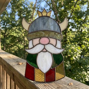 May include: Stained glass Viking ornament. It features a gray helmet, cream horns, and a gold crown. The face is pink with a white mustache and beard. The bottom is red, green, and gold. It hangs from a chain.