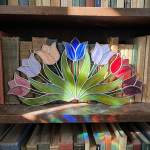 May include: A stained glass tulip design in shades of pink, purple, orange, blue, and red. The artwork is displayed on a wooden shelf in front of a collection of books. The piece is approximately 30 cm wide.