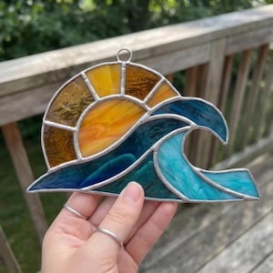 May include: A stained glass suncatcher featuring a semi-circular yellow and amber sun rising behind a stylized ocean wave. The wave is composed of various shades of blue and teal glass pieces held together by silver metal solder lines.