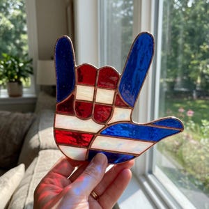 Könnte beinhalten: Eine Glasmalerei-Hand im "Ich liebe dich"-Zeichen, mit roten, weißen und blauen Farben. Das Design enthält Streifen und Quadrate mit kupferfarbenen Rändern. Das Stück wird vor einem Fenster gehalten und fängt das Licht ein.