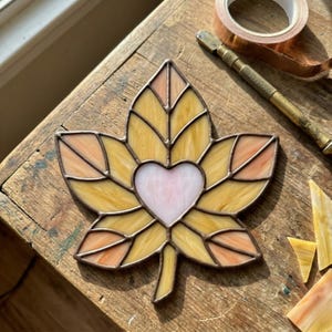 May include: Stained glass leaf with a heart-shaped center. The leaf features shades of yellow, orange, and pink glass, outlined with a copper-colored metal. The leaf is approximately 5 inches wide and is likely a decorative item.