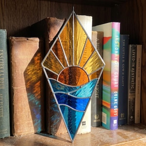 Op de afbeelding: Een ruitvormige glas-in-lood zonnevanger met een zonsopgangontwerp. Het bovenste gedeelte toont gele en oranje glazen stralen boven een donkeroranje zon, terwijl het onderste gedeelte blauwe en lichtblauwe golvende waterpatronen weergeeft.