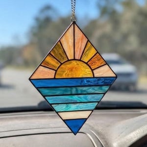Könnte beinhalten: Raute-förmiger Glas-Suncatcher. Das Design zeigt einen Sonnenuntergang über dem Meer, mit Gelb-, Orange- und Blautönen. Der Suncatcher hängt an einer Kette.