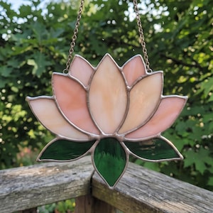 Op de afbeelding: Een glas-in-lood zonnevanger in de vorm van een lotusbloem met lichtroze bloemblaadjes en donkergroene bladeren aan de basis. De afzonderlijke glasstukken zijn verbonden door dunne metalen soldeerlijnen en het object hangt aan een fijne metalen ketting.