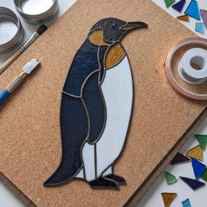 Puede incluir: Una figura de pingüino de vidriera con piezas de vidrio azul oscuro, blanco y dorado unidas por líneas de soldadura de metal oscuro. El pingüino tiene un detalle de ojo de vidrio azul y está junto a herramientas de metal y restos de vidrio.