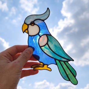 May include: A stained glass suncatcher shaped like a cockatiel bird. The bird features iridescent blue and teal glass panels, a white cheek patch, a yellow beak, and yellow feet, all outlined with dark metal solder lines against a cloudy sky.